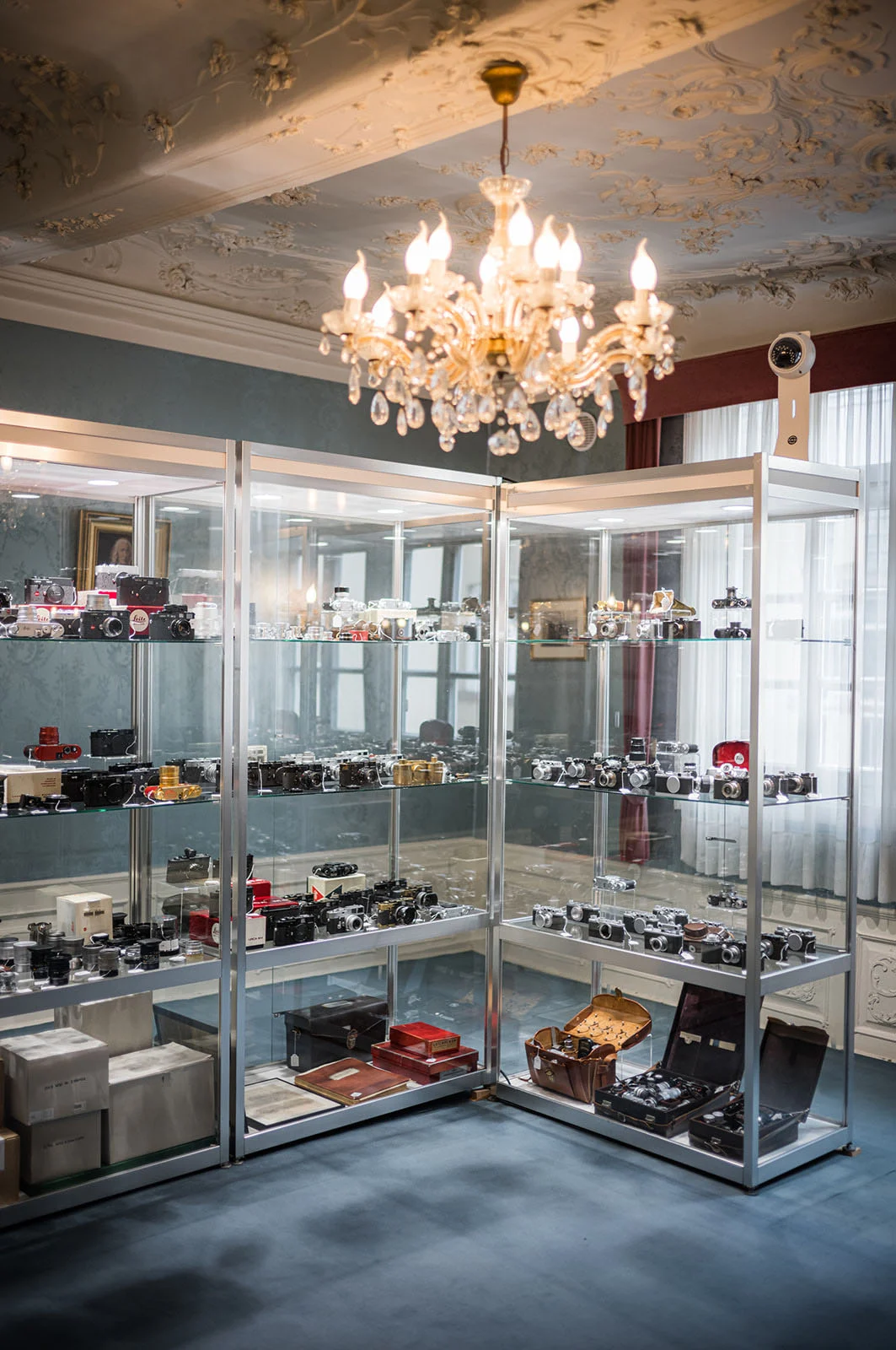 1923 Üretimi Leica 0-Series, 4,25 Milyon Euroya Satıldı 29 Three glass display cases filled with vintage cameras and accessories stand in a room with a crystal chandelier and ornate ceiling.