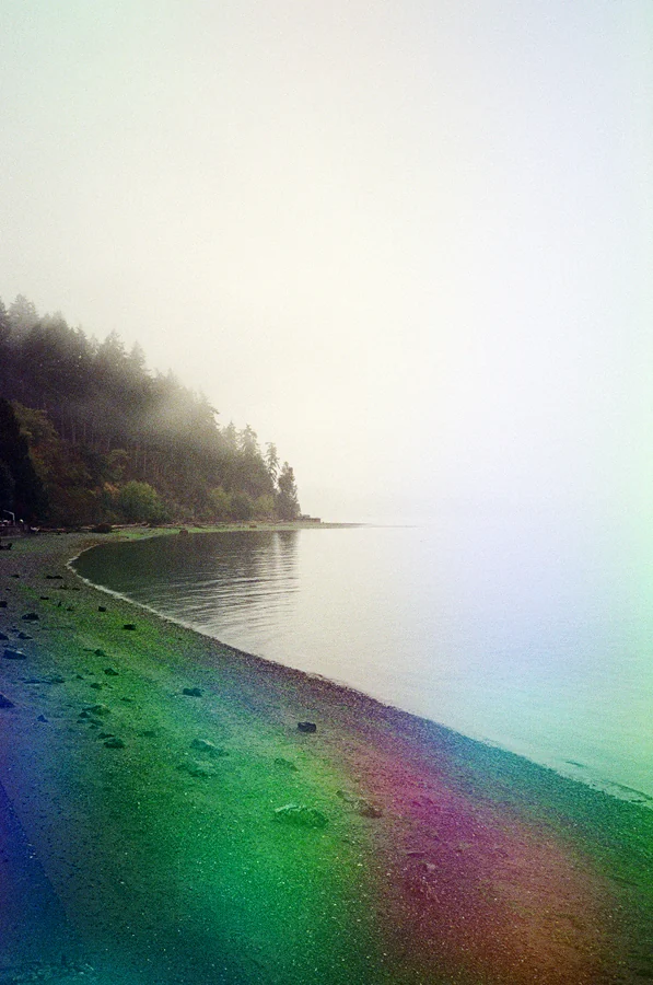 Psychedelic Blues: Analog Fotoğrafçılıkta Renk, Işık ve Rastlantının Peşinde 24 Sakin suyun kıyısında kıvrılarak uzanan sisli bir sahil ve sol tarafta ormanlık alan. Fotoğrafta gökkuşağı renklerinde ışık efektleri görülüyor.