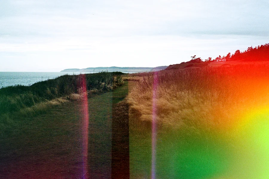 Psychedelic Blues: Analog Fotoğrafçılıkta Renk, Işık ve Rastlantının Peşinde 23 Bulutlu gökyüzü altında kıyı kayalıkları boyunca uzanan çimenli bir patika. Görüntünün üzerinde belirgin renkli ışık sızmaları ve ton bozulmaları bulunuyor.