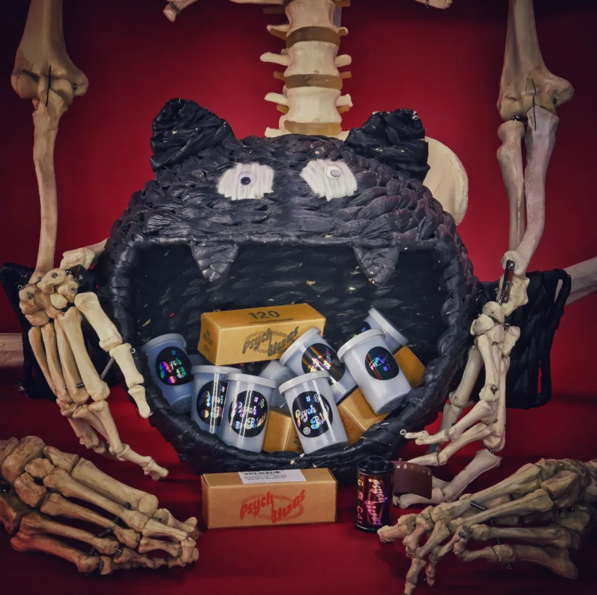 A skeleton holds a large, black, woven cat-shaped basket filled with several film canisters and boxes labeled "Psych Blues" against a red background.