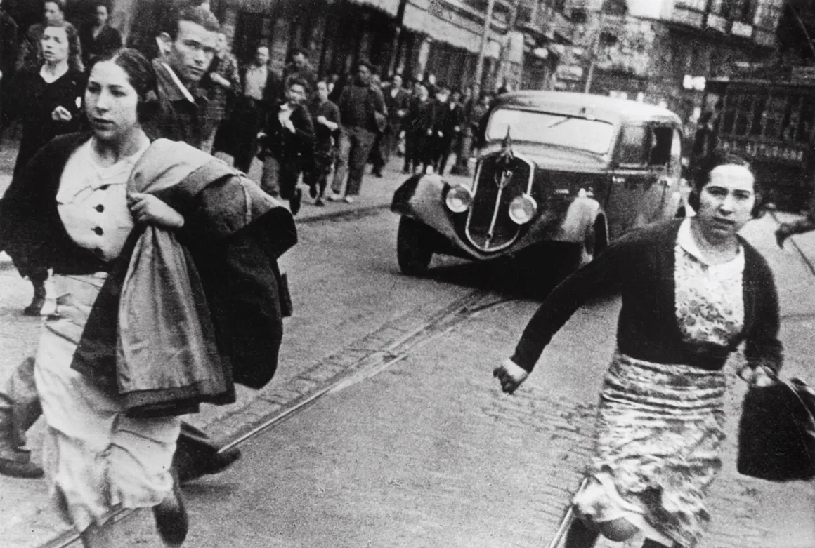 Robert Capa’nın Death in the Making Kitabı 88 Yıl Sonra Yeniden Basılıyor
İki kadın hava saldırısı sireni sırasında Arnavut kaldırımlı bir sokakta kameraya doğru koşuyor; arka planda eski bir otomobil ve başka yayalar görülüyor.