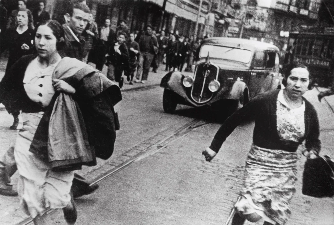 Two women run across a cobblestone street toward the camera, while a vintage car and other pedestrians move in the background.