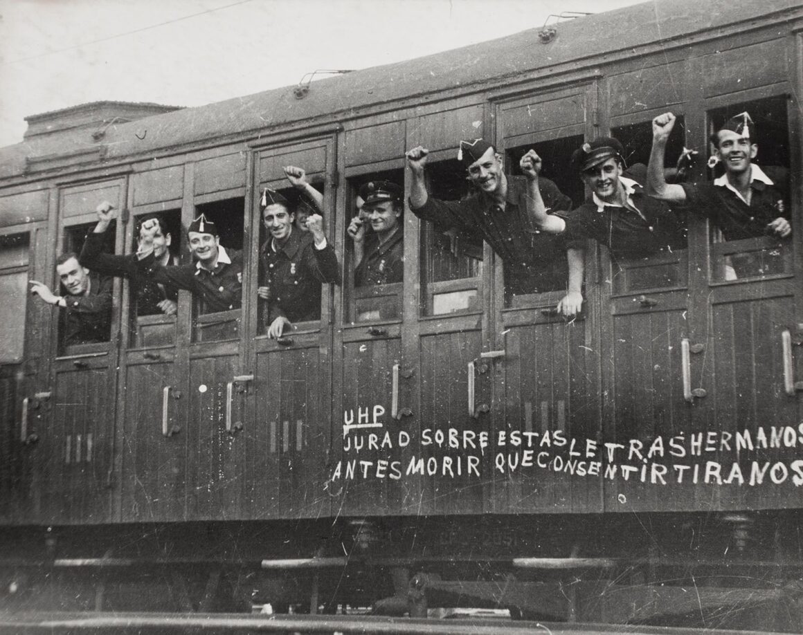 Robert Capa’nın Death in the Making Kitabı 88 Yıl Sonra Yeniden Basılıyor 20 Koyu renk üniformalı ve şapkalı bir grup erkek, ahşap tren vagonunun pencerelerinden dışarı uzanarak yumruklarını havaya kaldırıyor.
