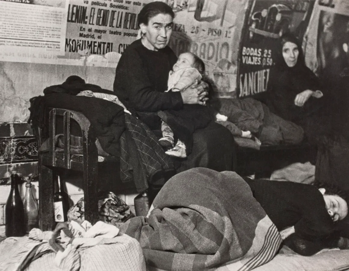 Robert Capa’nın Death in the Making Kitabı 88 Yıl Sonra Yeniden Basılıyor 22 Bir kadın bebeğini kucağında tutarak duvarlarında afişler bulunan bir odada oturuyor; yakınında başka bir kişi yerde uyuyor.