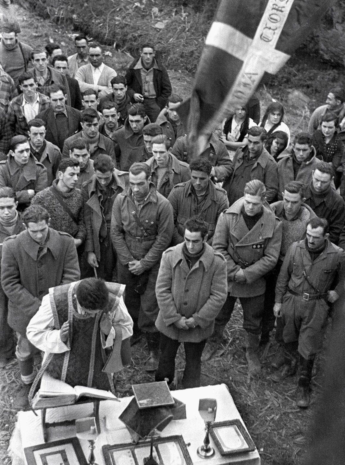 Robert Capa’nın Death in the Making Kitabı 88 Yıl Sonra Yeniden Basılıyor 24 Bir rahip açık havada askerî kıyafetli kalabalık bir erkek grubu için dini ayin gerçekleştiriyor; arka planda bir bayrak görülüyor.