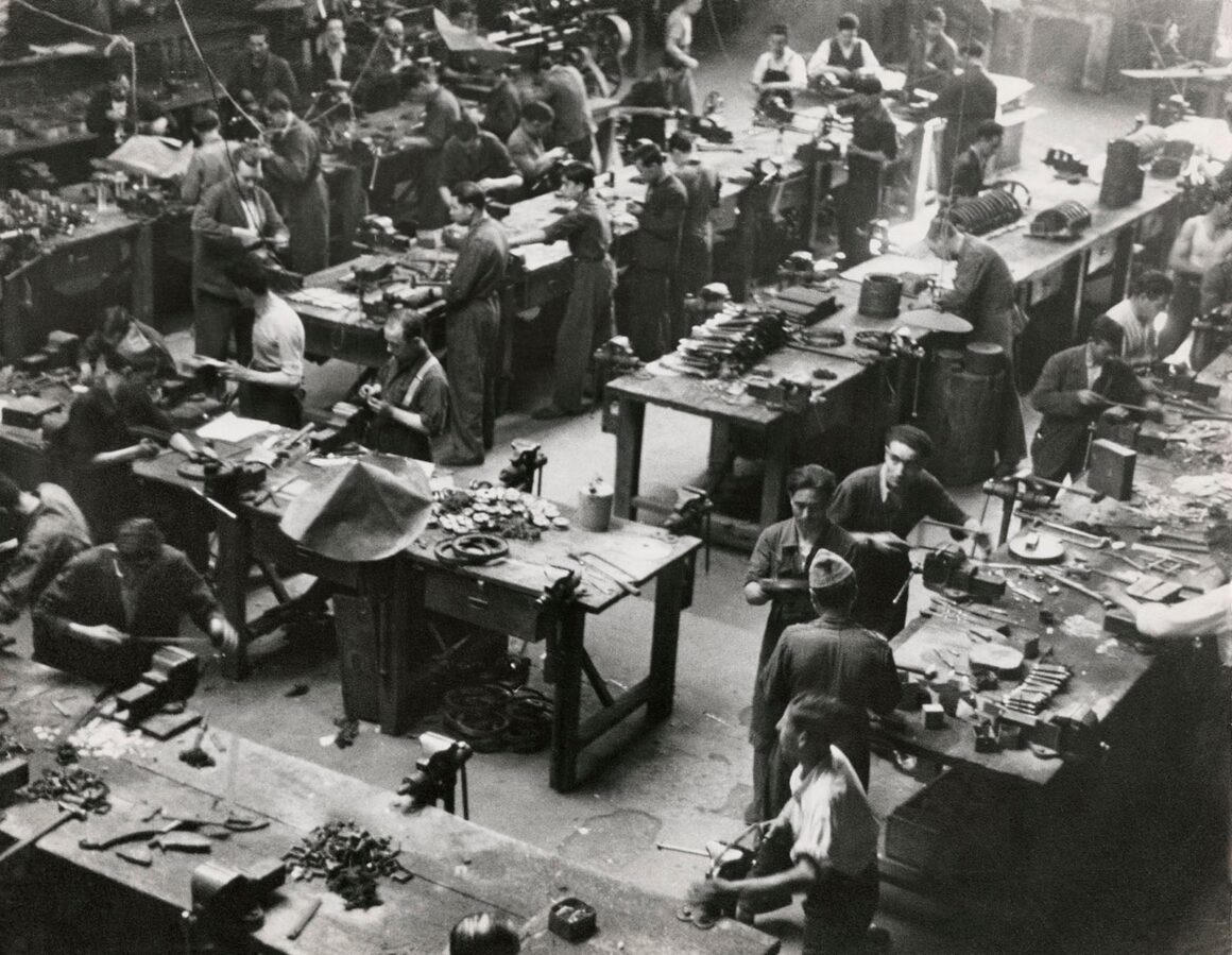 Robert Capa’nın Death in the Making Kitabı 88 Yıl Sonra Yeniden Basılıyor 25 Çok sayıda erkeğin mengene ve el aletleri bulunan ahşap tezgâhlarda çalıştığı kalabalık bir endüstriyel atölyenin yüksek açıdan görünümü.