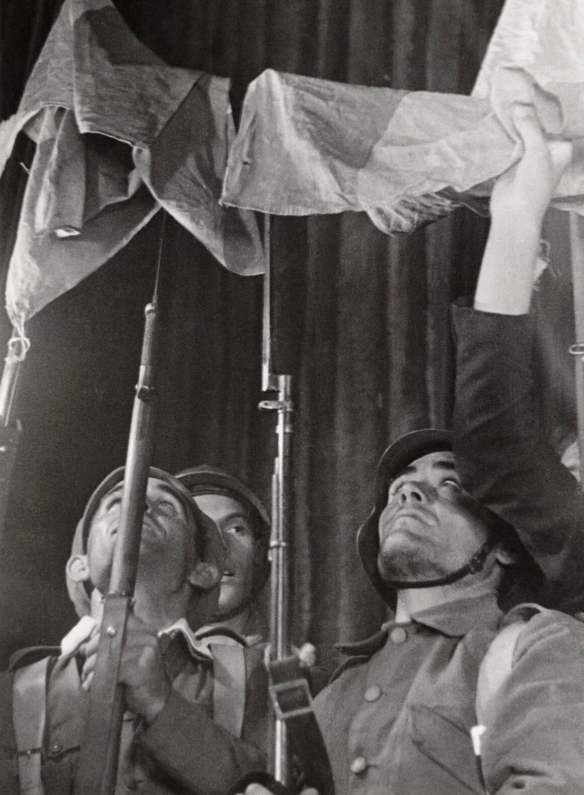 Robert Capa’nın Death in the Making Kitabı 88 Yıl Sonra Yeniden Basılıyor 26 Miğferli üç asker, tüfekler ve kumaş bir bayrak tutarken yukarı doğru bakıyor.