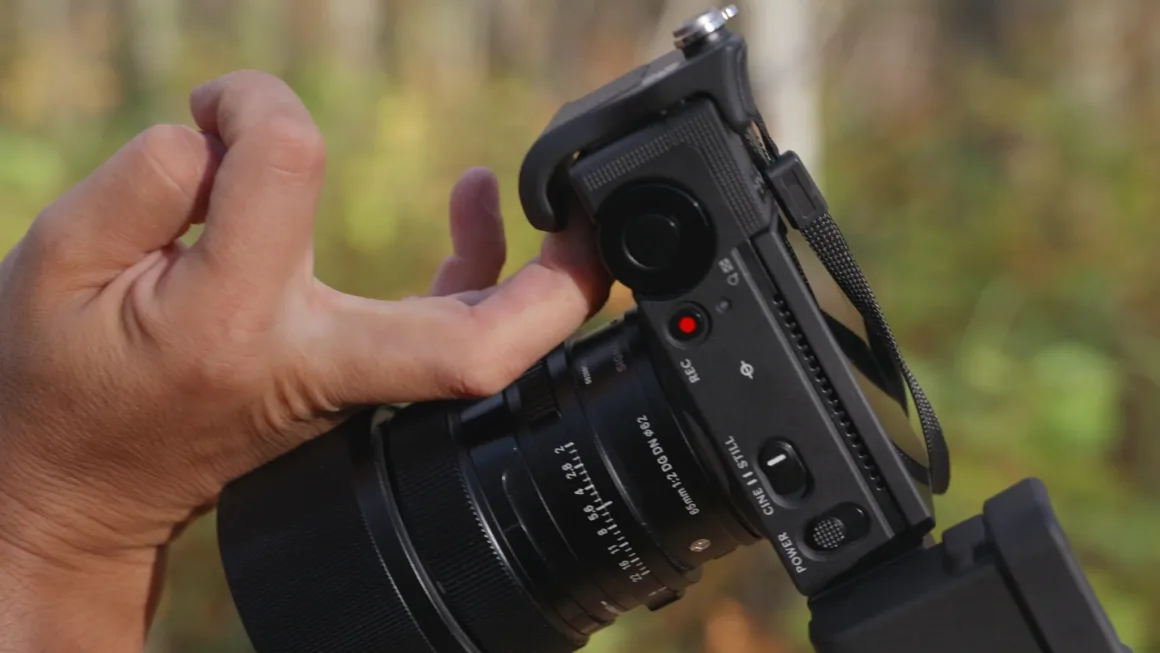Sigma FP2 İncelemesi: Tuhaf Tasarım, Güçlü Hibrit Performans 26 Bir kişinin eli Sigma 65mm objektif takılı siyah dijital kamerayı tutuyor; üst panelde kırmızı kayıt düğmesi ve güç anahtarı görülüyor.
