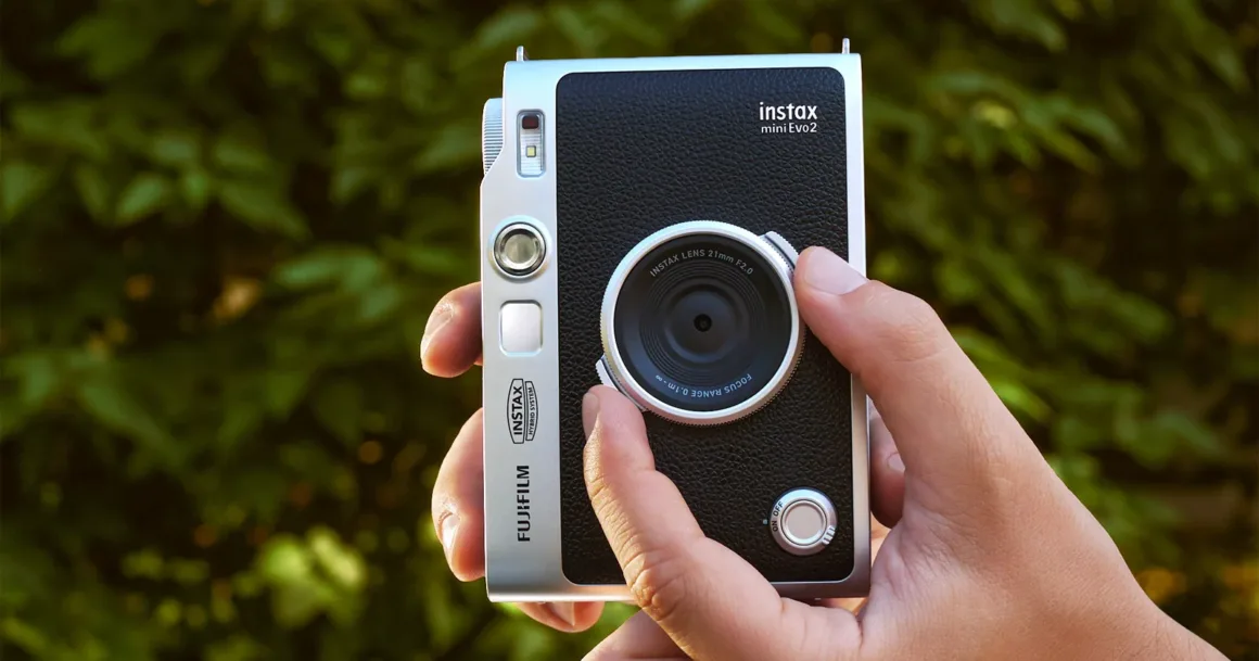 Bir el, bulanık yeşil yapraklı arka plan önünde gümüş ve siyah Fujifilm Instax Mini Evo 2 fotoğraf makinesini tutuyor.