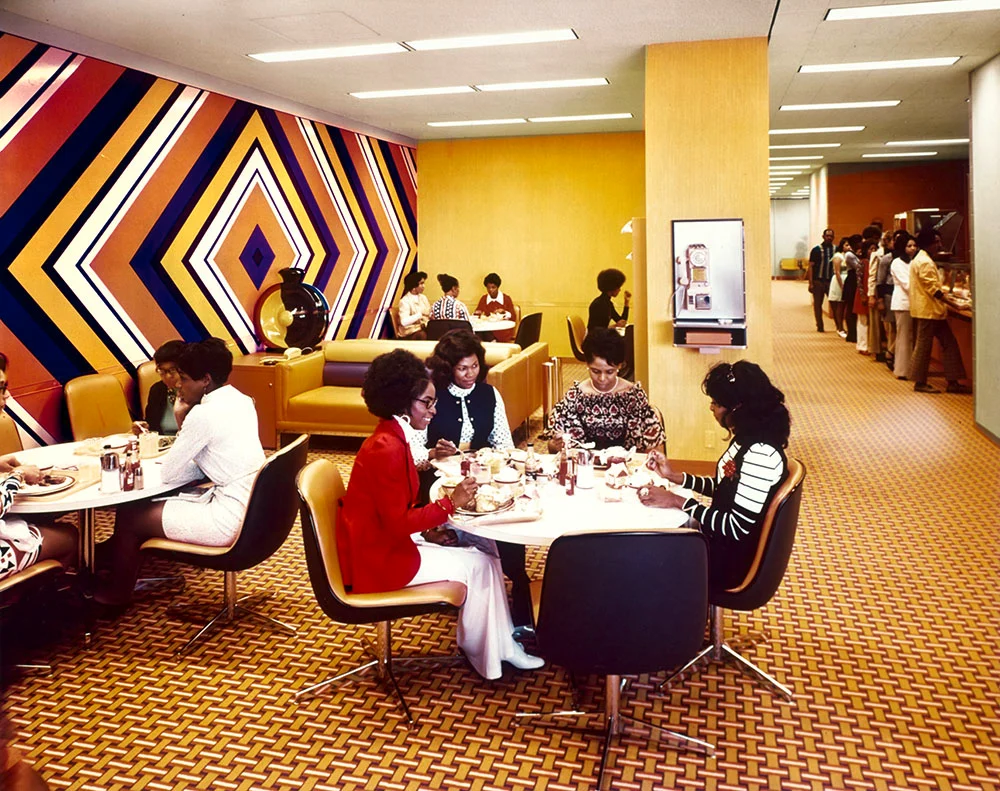 A group of women eat lunch at round tables in a brightly decorated cafeteria with a geometric wall mural and patterned carpet.