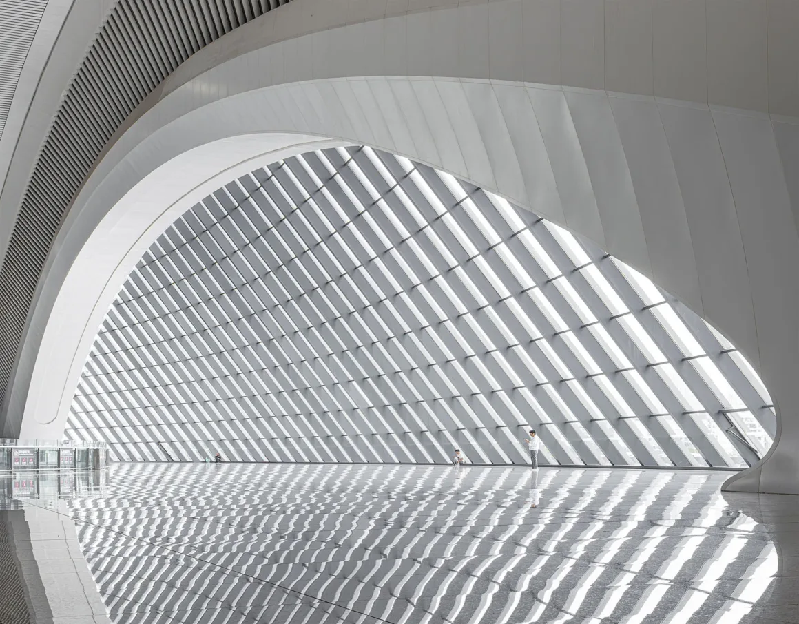 Architecture Photography MasterPrize 2026 Kazananları Açıklandı 30 Büyük kıvrımlı beyaz mimari yapı, çapraz ızgara desenli cam duvarı çerçeveliyor; yansımalı zeminde iki kişi duruyor.
