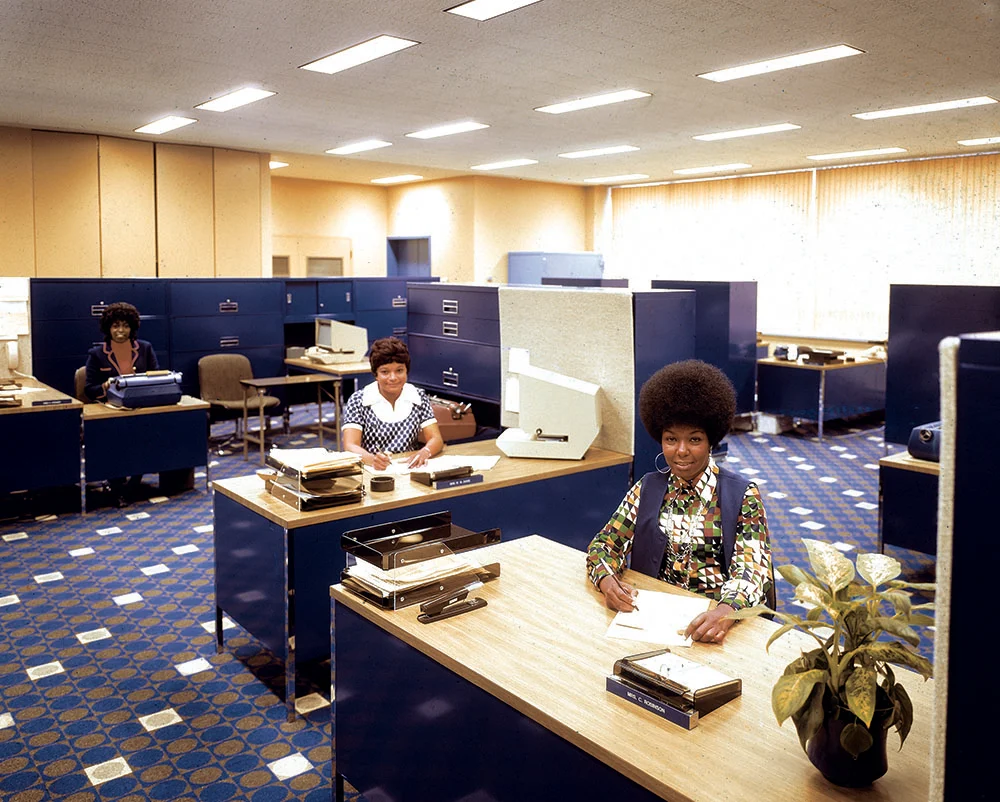 Three women work at desks in a modern office with blue filing cabinets and a patterned blue and white carpet.