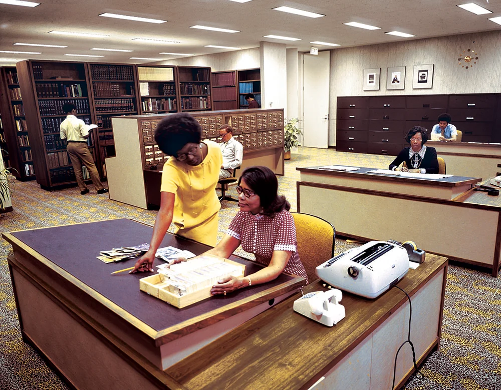 A library office scene with several people working at desks, including two women in the foreground reviewing index cards.
