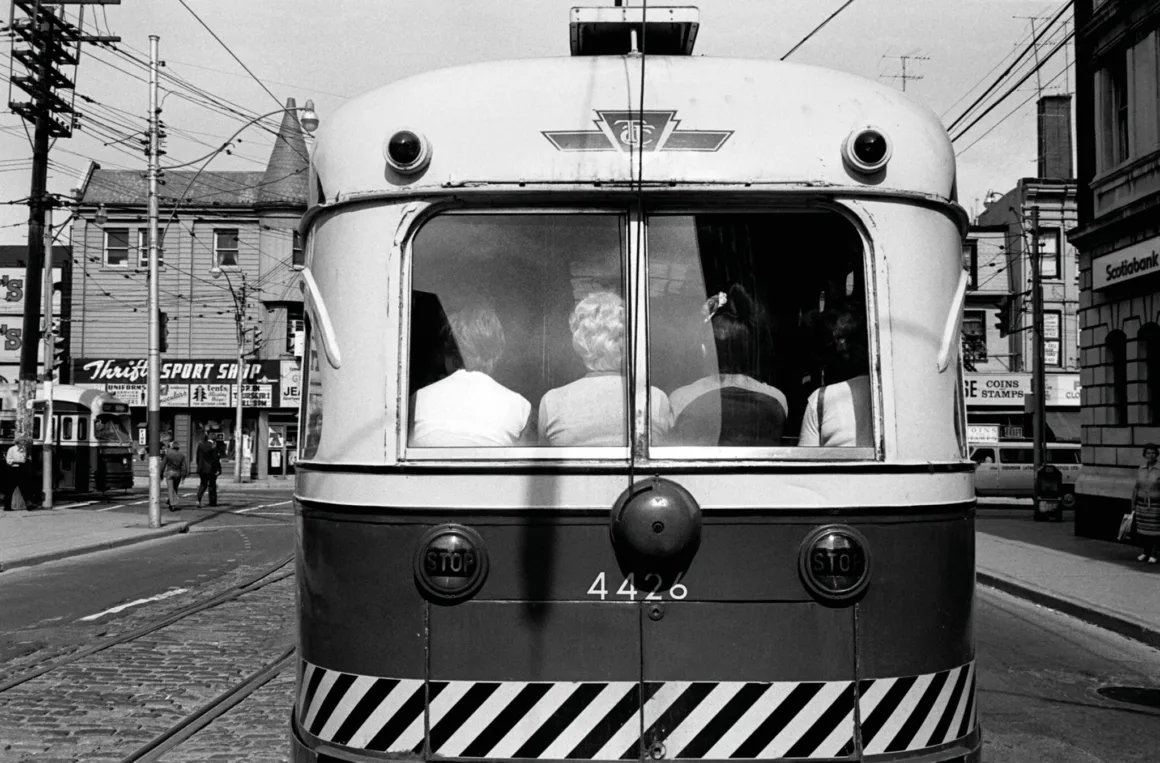 4426 numaralı Toronto Transit Commission tramvayının arka bölümünde içeride oturan dört yolcu görülürken, arka planda şehir dükkânları ve sokak tabelaları yer alıyor.