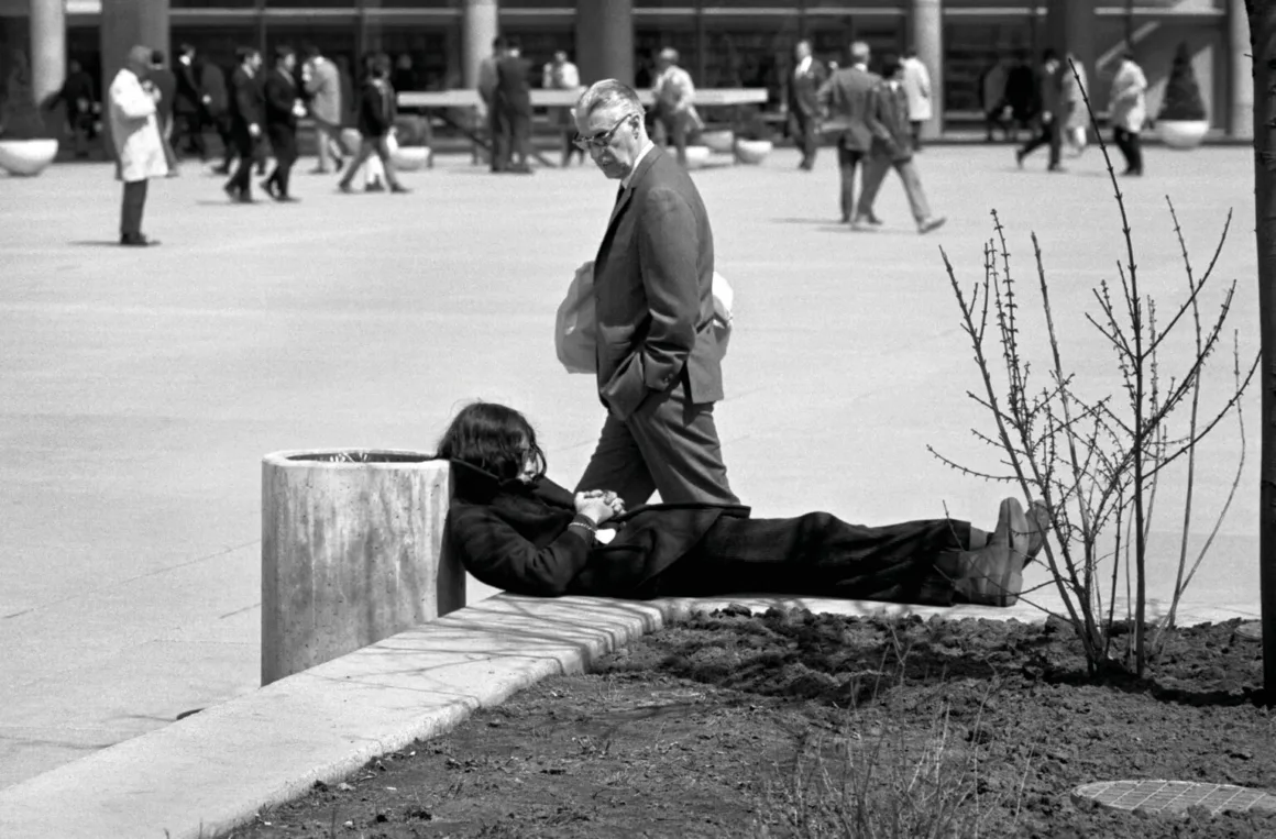 Brian Condron’ın Objektifinden 1970’lerin Toronto’su
Uzun koyu saçlı bir kişi çöp kutusunun yanında beton bir çıkıntının üzerinde uzanıp dinlenirken, arka planda takım elbiseli bir adam yürüyerek geçiyor.