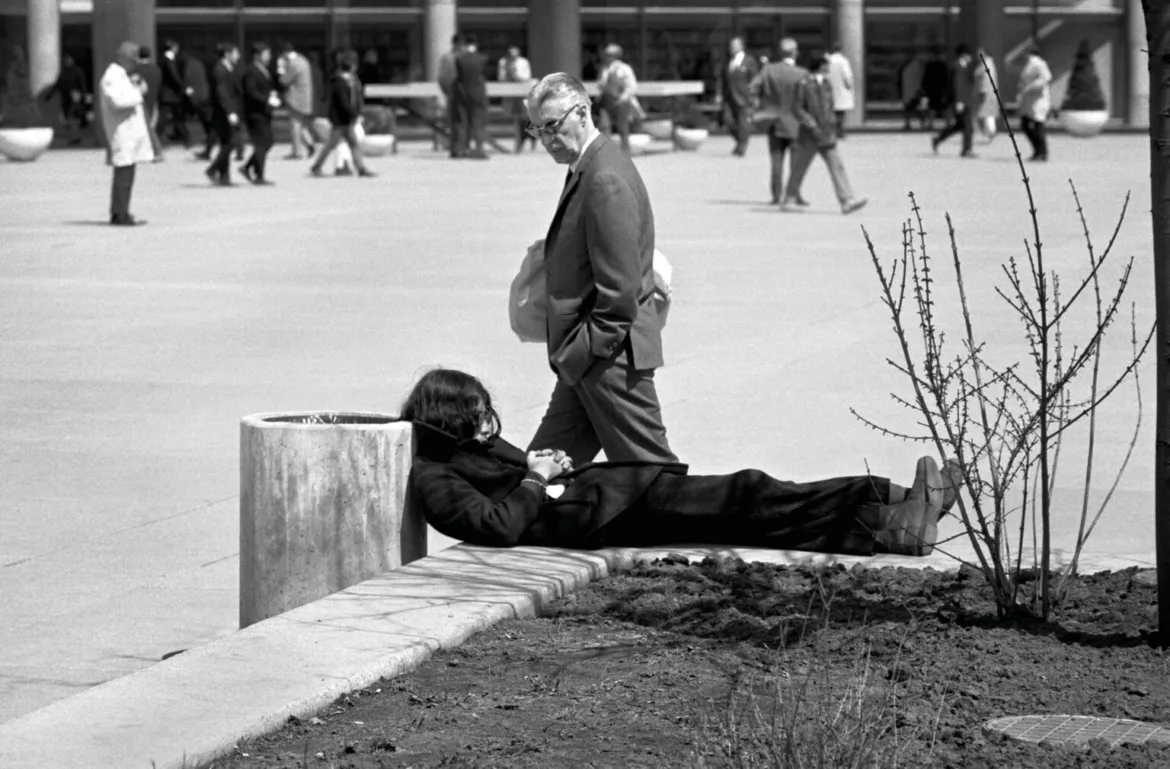 A person with long dark hair lies resting on a concrete ledge next to a trash bin while a man in a suit walks past in the background.