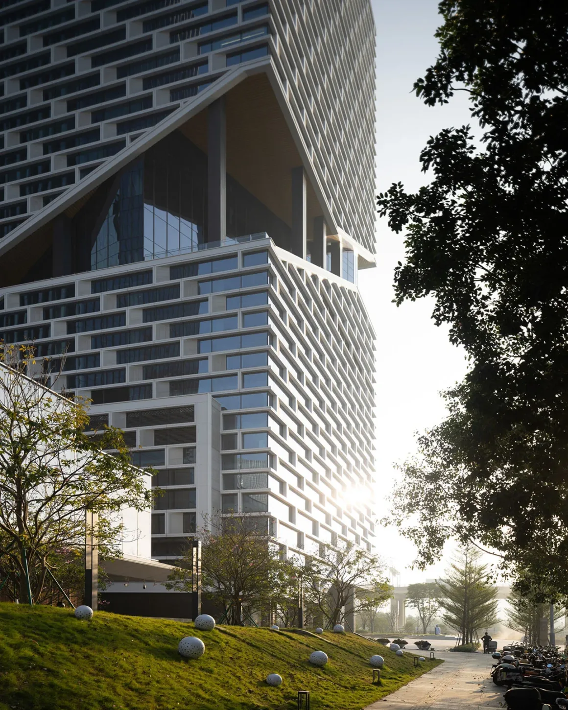 Architecture Photography MasterPrize 2026 Kazananları Açıklandı 31 Belirgin geometrik cephesi ve büyük açık hava boşluğuyla modern gökdelen, dekoratif kürelerin ve yürüyüş yolunun bulunduğu çimenlik eğimin üzerinde yükseliyor.