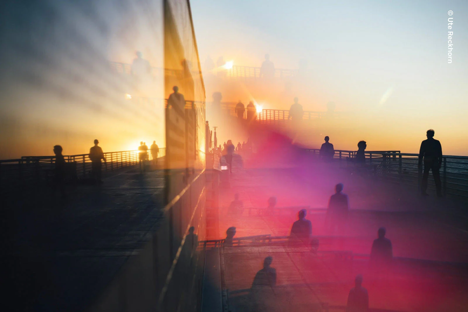Lensbaby Omni Color II ve Omni Crystal II ile Çekim Sırasında Yaratıcı Efektler 23 A double-exposure photograph shows silhouettes of people walking on a pier at sunset, with a prominent pink light flare overlaying the scene.