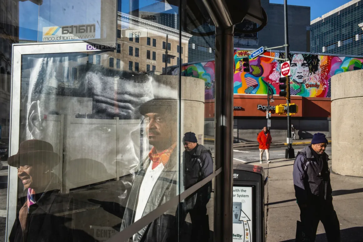 Alex Webb’in Walking Blues’u: Amerika Sokaklarında Renk, Ritim ve Rastlantı
Bir otobüs durağının cam panelinde şapkalı bir adamın yansıması görülürken yayalar şehir sokağındaki renkli bir duvar resminin önünden geçiyor.