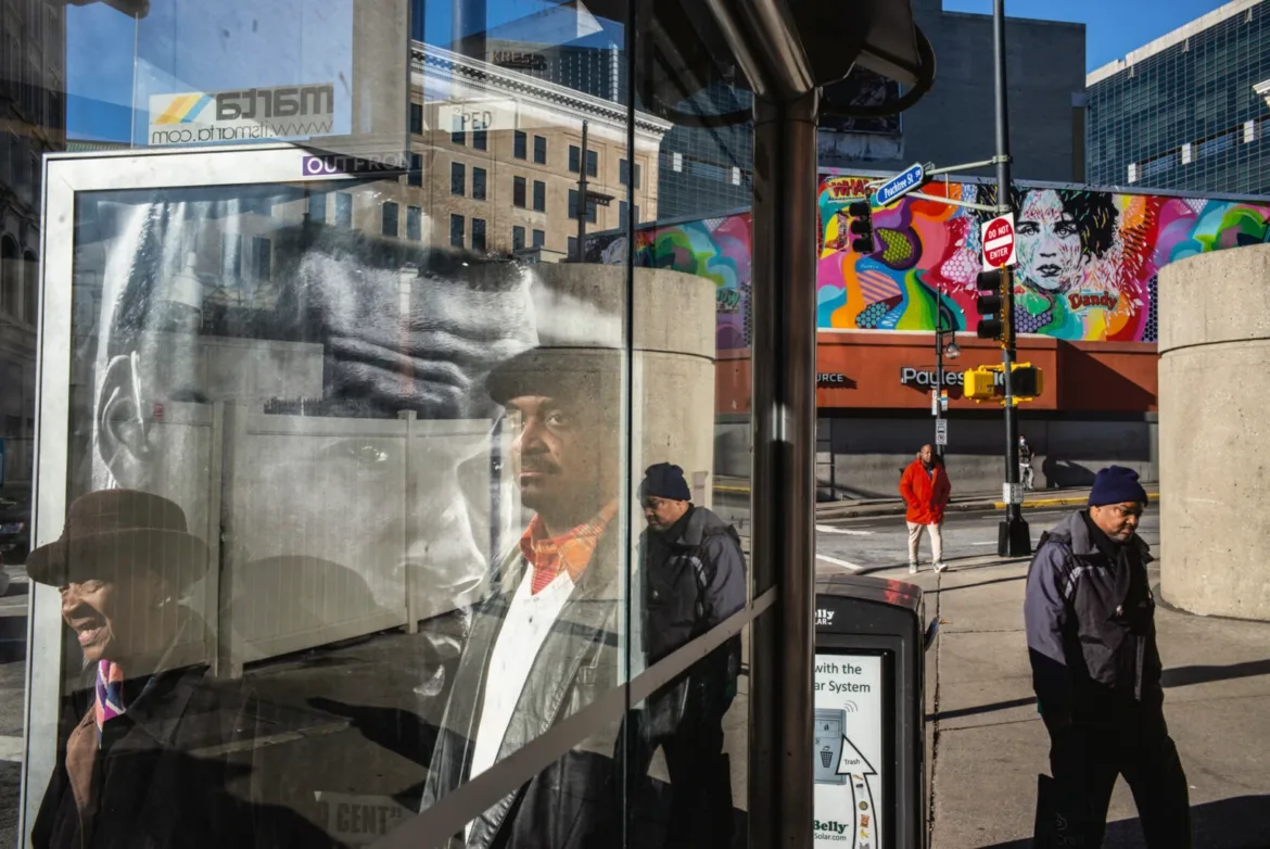 A bus shelter glass panel reflects a man in a hat while pedestrians walk past a colorful mural on a city street.