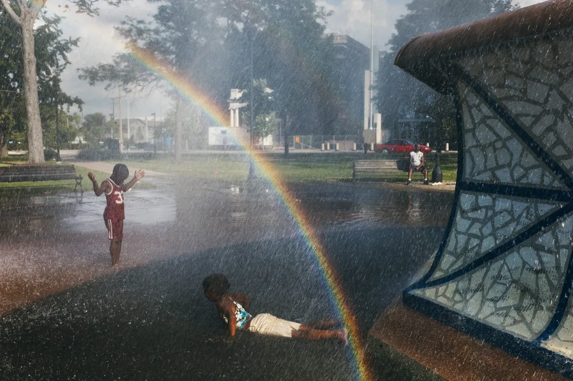 Alex Webb’in Walking Blues’u: Amerika Sokaklarında Renk, Ritim ve Rastlantı 20 İki çocuk park çeşmesinin püskürttüğü suyun içinde oynarken gökkuşağı oluşuyor; arka planda bir yetişkin bankta oturuyor.