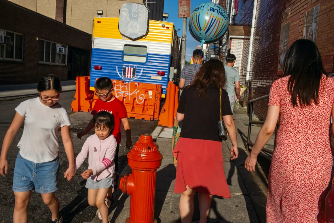 Alex Webb’in Walking Blues’u: Amerika Sokaklarında Renk, Ritim ve Rastlantı 22 Bir grup insan, parlak turuncu yangın musluğunun ve inşaat bariyerlerinin arkasında duran büyük mavi-sarı treylerin önünden kaldırım boyunca yürüyor.