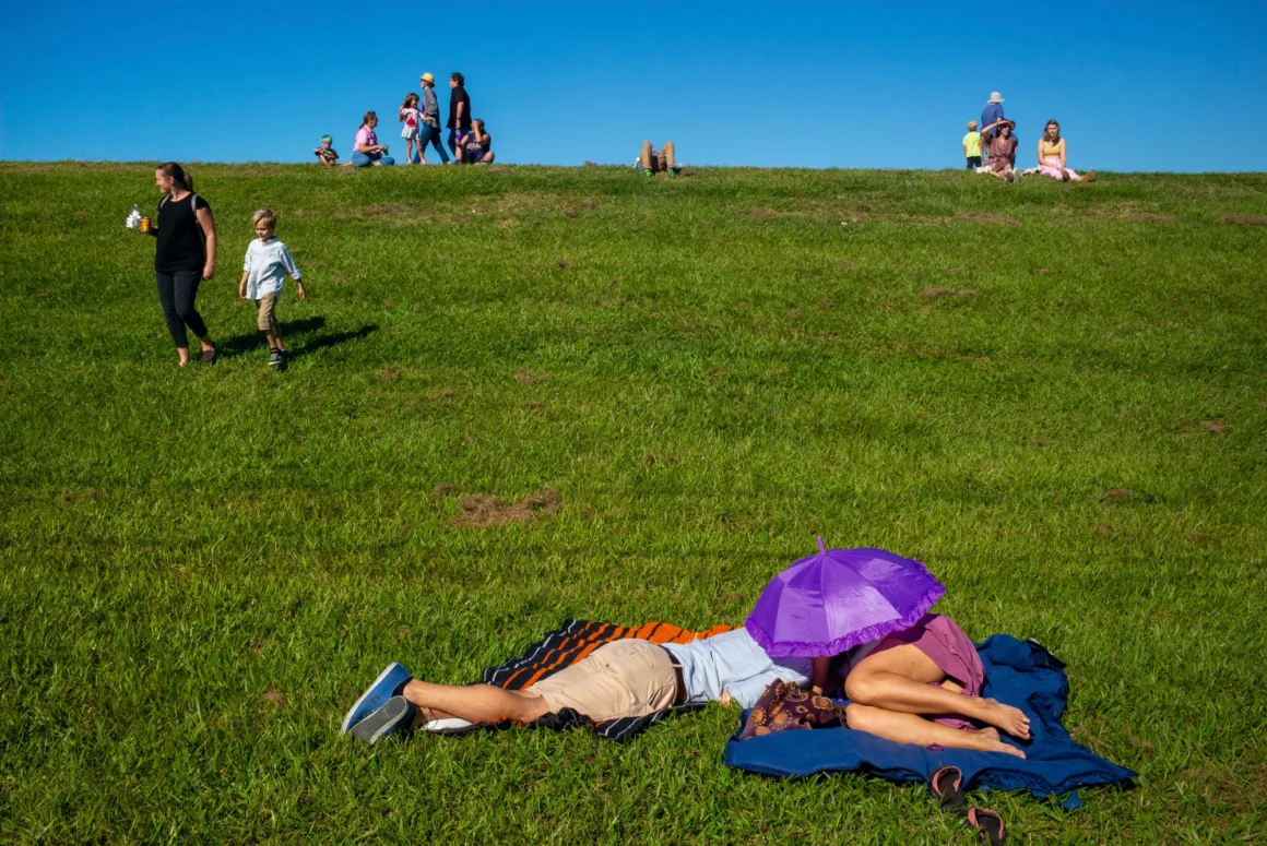 Alex Webb’in Walking Blues’u: Amerika Sokaklarında Renk, Ritim ve Rastlantı 23 İnsanlar parlak mavi gökyüzü altında çimenlik bir tepede dinlenirken ön planda mor bir şemsiyenin altında uzanan bir çift görülüyor.