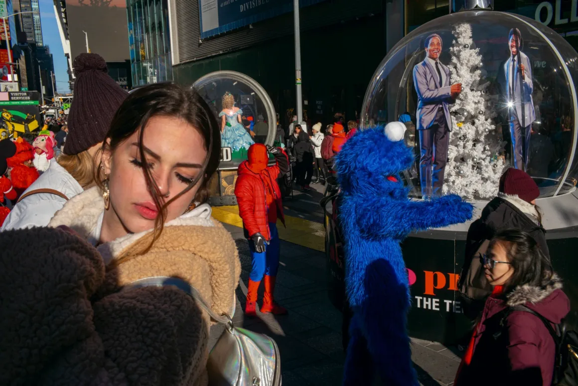 Alex Webb’in Walking Blues’u: Amerika Sokaklarında Renk, Ritim ve Rastlantı 24 Bej polar ceket giyen genç bir kadın, kalabalık şehir sokağında Cookie Monster kostümlü ve Spider-Man kıyafetli kişilerin yakınında duruyor.