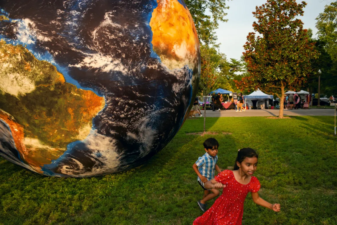 Alex Webb’in Walking Blues’u: Amerika Sokaklarında Renk, Ritim ve Rastlantı 25 Kırmızı elbiseli genç bir kız ve mavi kareli gömlekli bir erkek çocuk, büyük şişme dünya küresinin yanındaki çimenlik parkta koşuyor.