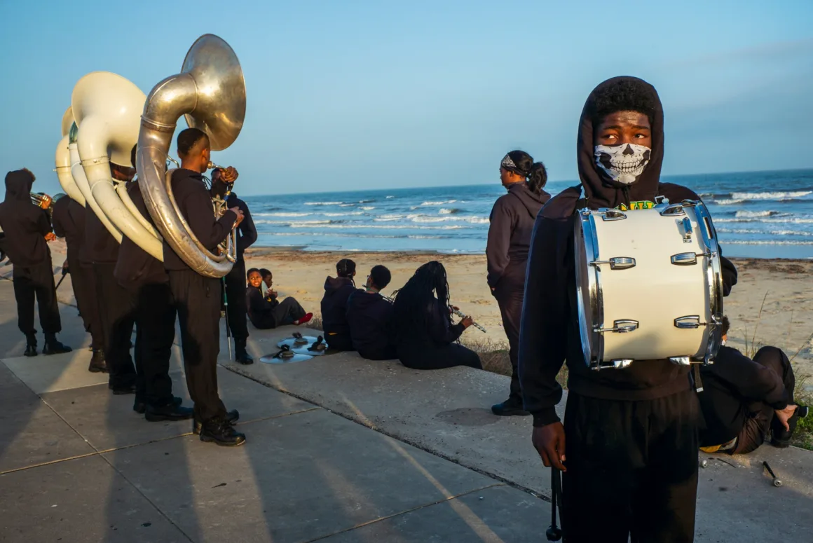 Alex Webb’in Walking Blues’u: Amerika Sokaklarında Renk, Ritim ve Rastlantı 28 Kafatası desenli yüz maskesi takan ve büyük davul taşıyan genç bir kişi ön planda dururken siyah kapüşonlu diğer bando üyeleri deniz kenarındaki beton yolda dinleniyor.