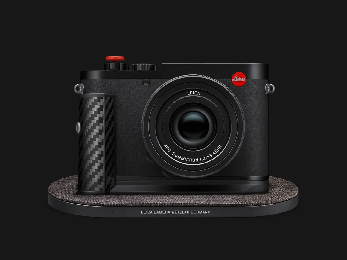 Leica Q3 43 Formula 1 Edition: Karbon Fiber Detaylı Özel Seri 29 Karbon fiber tutma bölümüne ve kırmızı deklanşör düğmesine sahip siyah Leica fotoğraf makinesi, dokulu oval biçimli kablosuz şarj yüzeyi üzerinde duruyor.