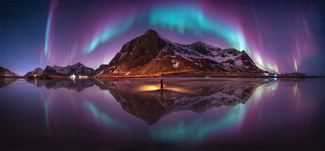 Epson International Pano Awards 2026 Kazananları Açıklandı 21 Bir kişi gece vakti yansıtıcı kıyıda dururken büyük bir dağın üzerinde canlı ve kavisli kuzey ışıkları yükseliyor.