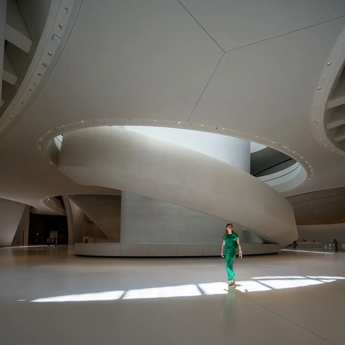 Architecture Photography MasterPrize 2026 Kazananları Açıklandı 32 Yeşil kıyafetli bir kişi, büyük kıvrımlı rampa ve dairesel tavan açıklığının bulunduğu geniş, minimalist ve beyaz zeminli salonda yürüyor.