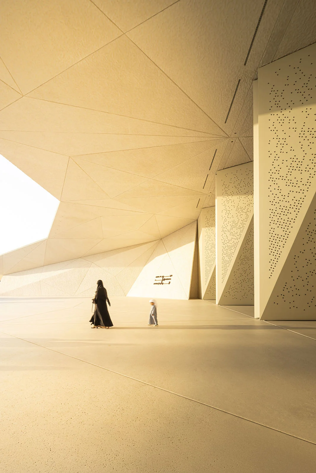 Architecture Photography MasterPrize 2026 Kazananları Açıklandı 33 Siyah abaya giymiş bir kadın ile geleneksel kıyafetli küçük çocuk, büyük geometrik açık renkli mimari örtünün altındaki geniş minimalist meydanda yürüyor.