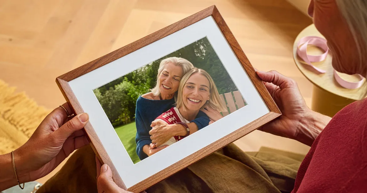 A person holds a framed photograph showing an older woman embracing a younger woman outdoors.