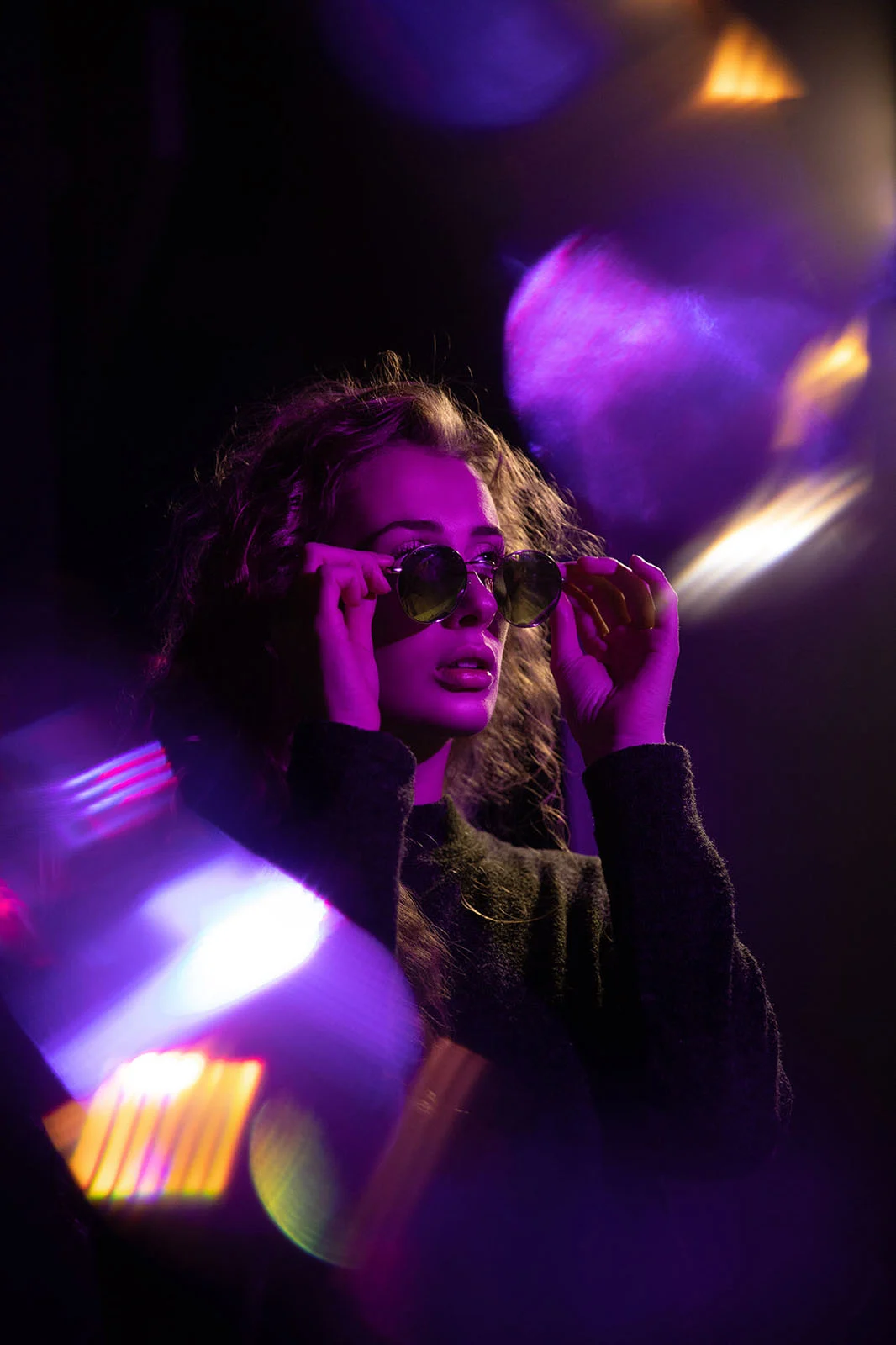 Lensbaby Omni Color II ve Omni Crystal II ile Çekim Sırasında Yaratıcı Efektler 20 A woman with curly hair wears round sunglasses while posing in a dark environment with vibrant purple and orange light flares.