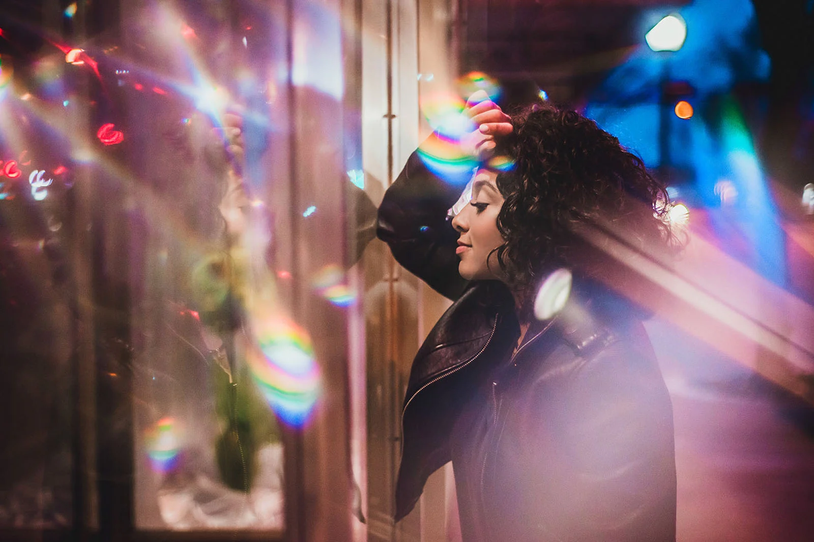 Lensbaby Omni Color II ve Omni Crystal II ile Çekim Sırasında Yaratıcı Efektler 25 A woman with curly hair and a leather jacket stands beside a glass storefront at night, surrounded by colorful lens flares.