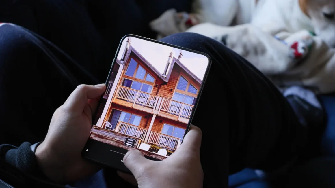 iPhone Duo İncelemesi: Etkileyici Tasarım, Kaçınılmaz Tavizler 25 Bir kişi, balkonlu ahşap bir kulübenin fotoğrafını gösteren katlanabilir akıllı telefonu tutuyor; arka planda bir köpek dinleniyor.