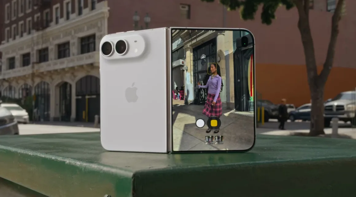 Apple iPhone Duo: İlk Katlanabilir iPhone 7,6 İnç Ekranla Geldi 30 Beyaz katlanabilir bir akıllı telefon yeşil bir yüzey üzerinde dik duruyor ve iç ekranında kamera arayüzü görülüyor.