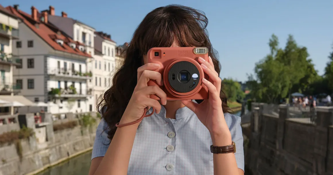 Fujifilm Instax Square 2: Yeni Tasarım, Yeni Mood Filtreleri 22 Uzun kahverengi saçlı bir kişi, terracotta renkli Instax Square 2 fotoğraf makinesini yüzüne doğru tutuyor; arka planda kanal ve binalar görülüyor.