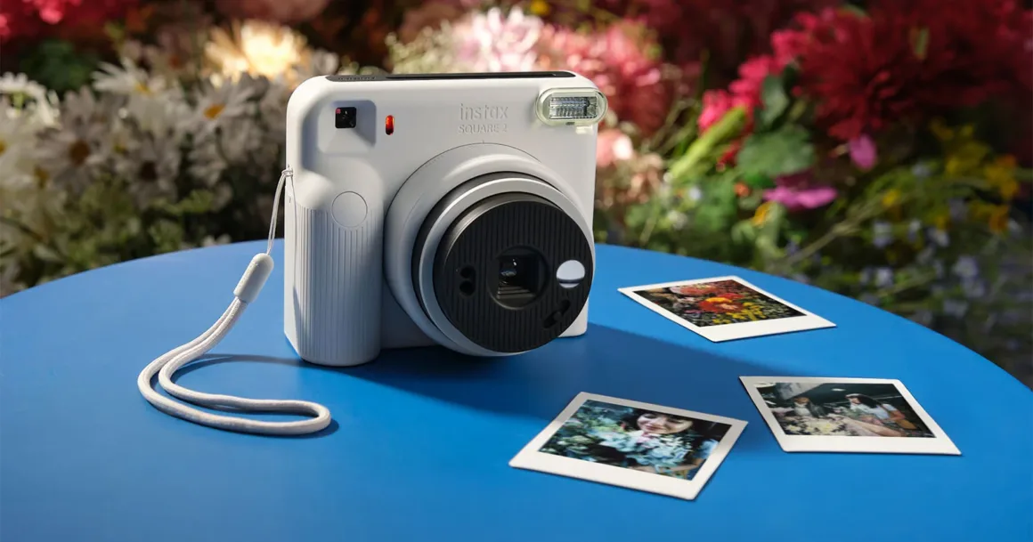 Fujifilm Instax Square 2: Yeni Tasarım, Yeni Mood Filtreleri 20 Beyaz Instax Square 2 anlık fotoğraf makinesi mavi bir masa üzerinde, yanında üç geliştirilmiş kare fotoğrafla birlikte duruyor; arka planda bulanık çiçekler görülüyor.