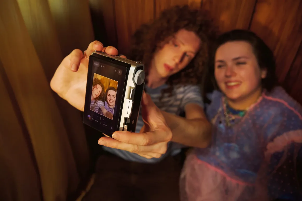 A person with curly hair holds a digital camera to take a selfie with a smiling woman beside them, with the camera screen displaying their live image.
