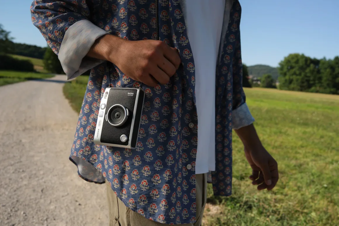 A person wearing a blue floral button-down shirt over a white t-shirt carries a silver and black Fujifilm Instax camera hanging from a strap.