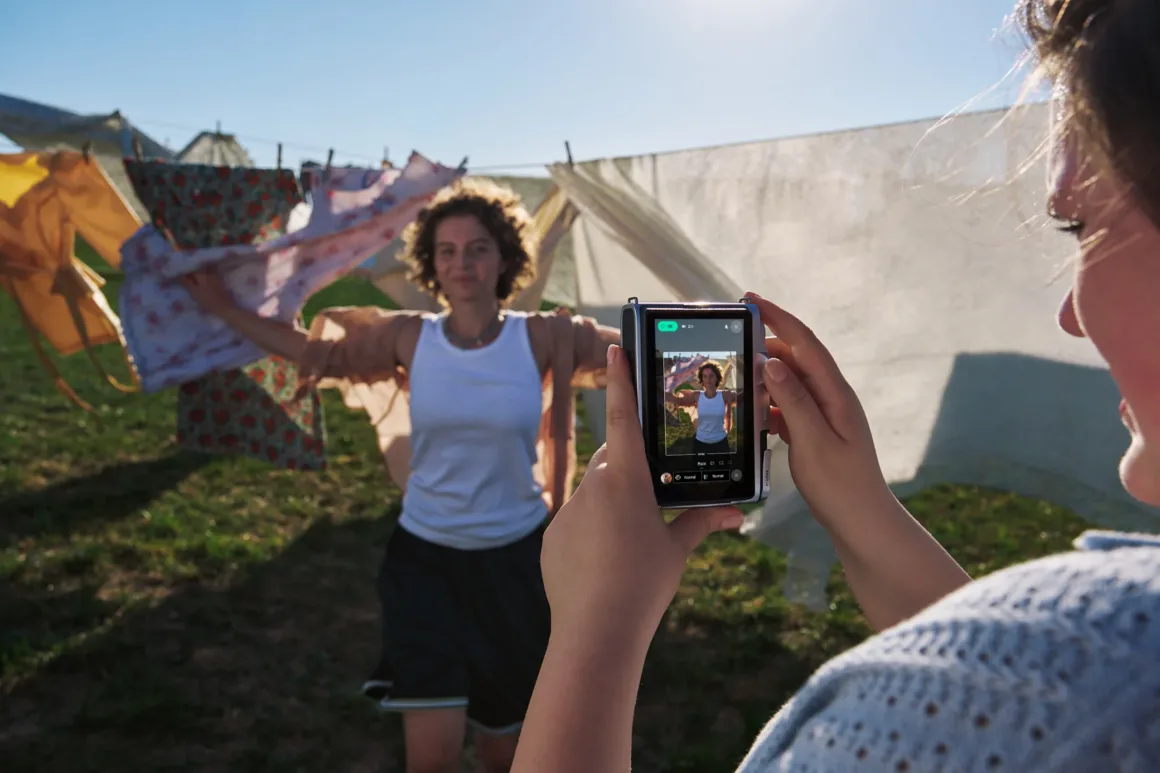 A person holds a digital camera to photograph another person standing in front of laundry hanging on a clothesline outdoors.