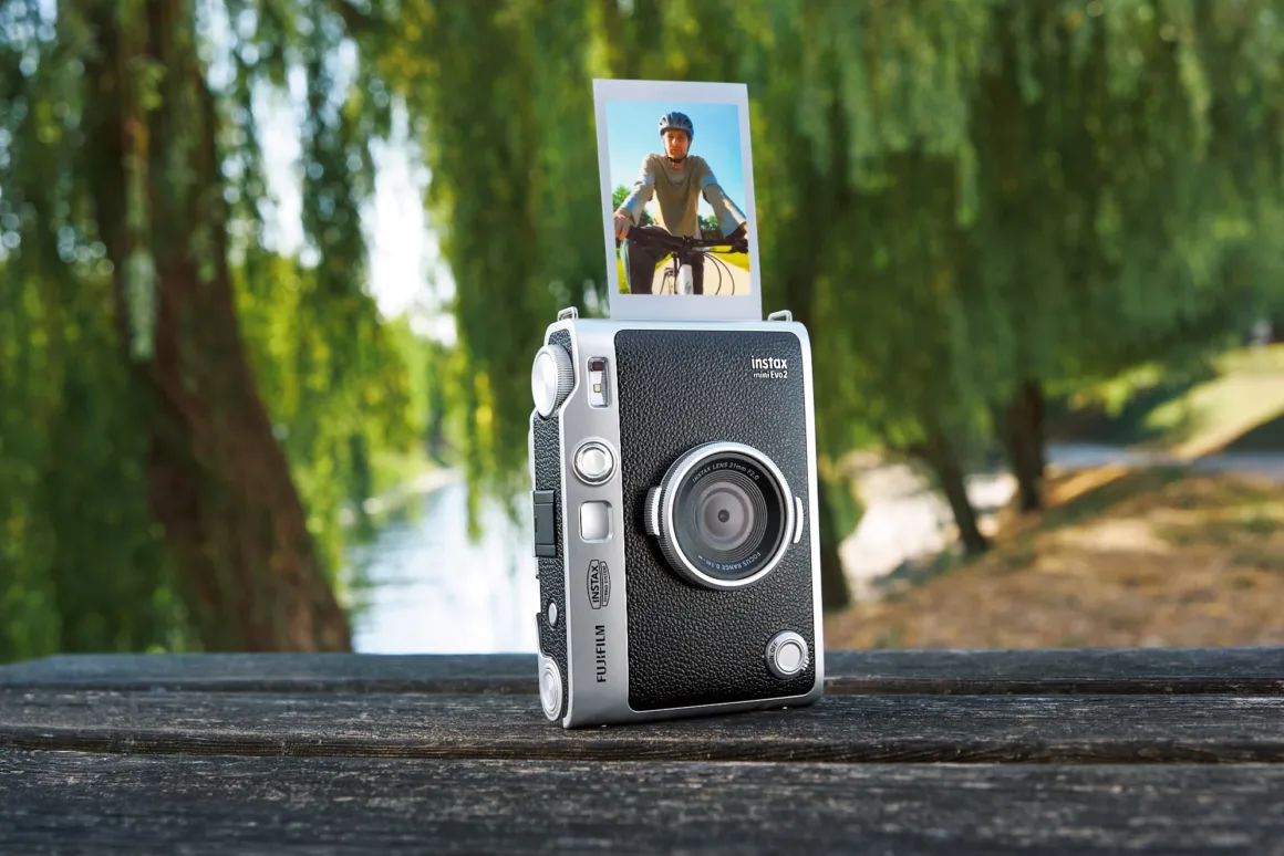 Fujifilm Instax Mini Evo fotoğraf makinesi dış mekânda ahşap yüzey üzerinde duruyor ve üst bölümünden bisikletçi fotoğrafının baskısı çıkıyor.