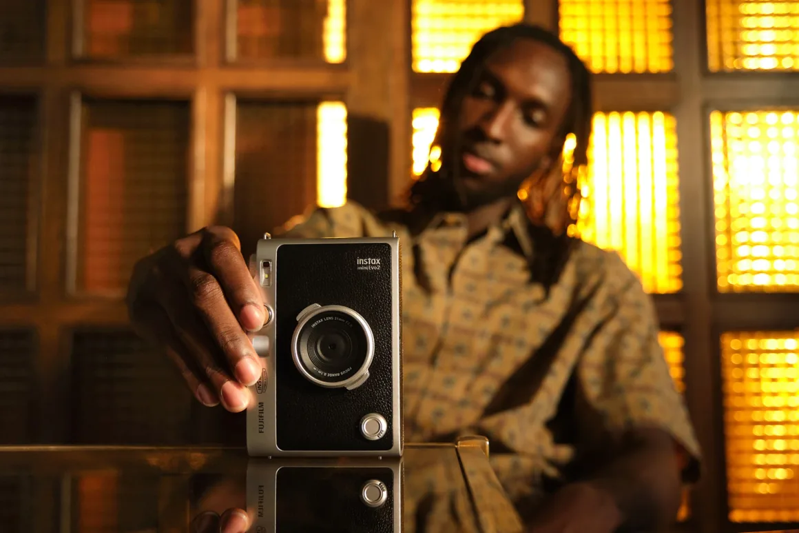 A person with dreadlocks sits behind a table, holding a silver and black Instax mini Evo 2 camera upright on the surface.