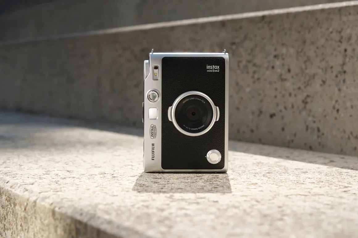 Gümüş ve siyah Fujifilm Instax Mini Evo 2 hibrit fotoğraf makinesi parlak güneş ışığında dokulu beton basamak üzerinde dik duruyor.