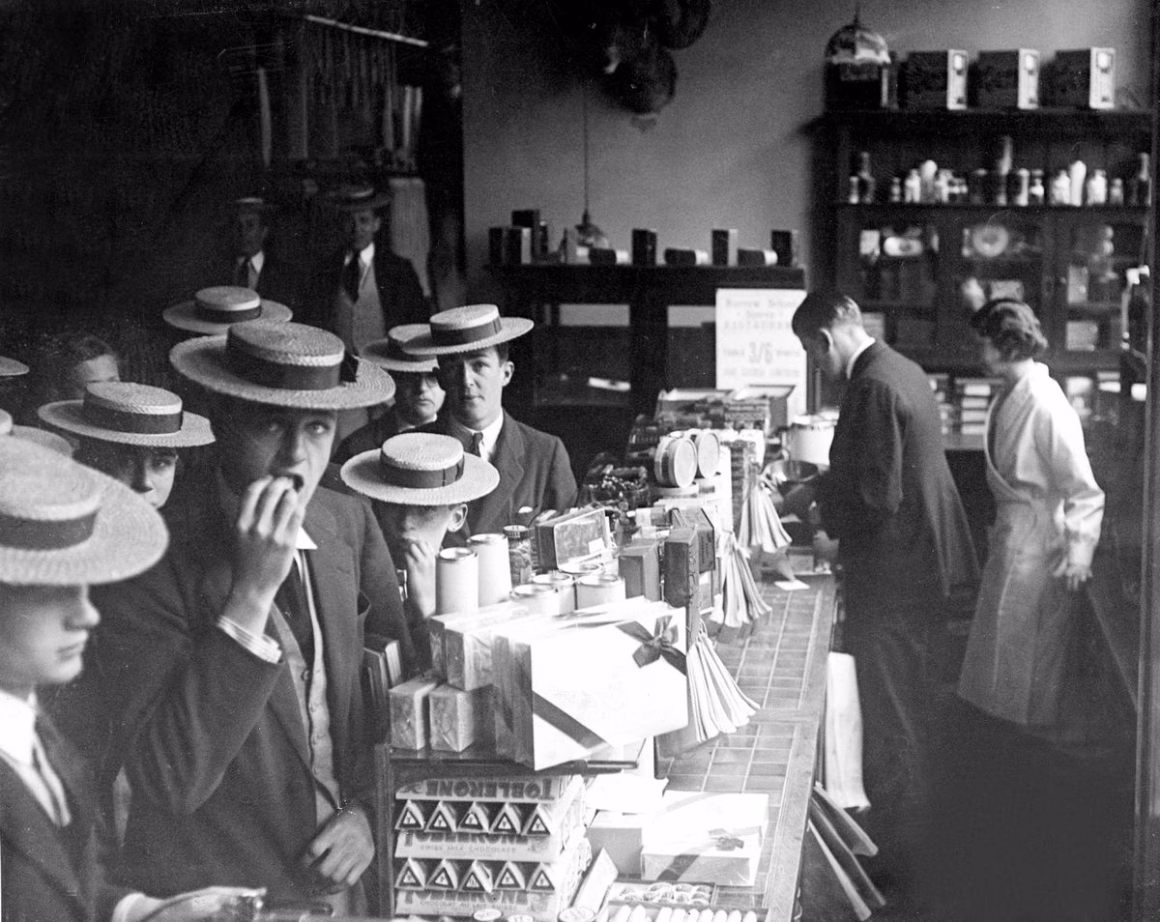 1930’ların Britanyası: Gündelik Hayatı Belgeleyen Unutulmuş Fotoğraflar 23 Hasır boater şapkalar takan öğrenciler, Toblerone ve şekerleme kutularıyla dolu dükkân tezgâhının önünde toplanmış; öğrencilerden biri ağzında yiyecekle doğrudan kameraya bakıyor.