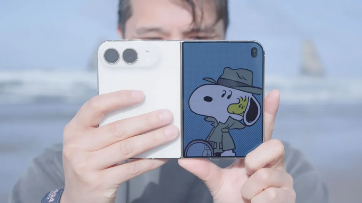 iPhone Duo İncelemesi: Etkileyici Tasarım, Kaçınılmaz Tavizler 31 Bir kişi, ekranında dedektif Snoopy animasyonu bulunan beyaz katlanabilir akıllı telefonu açık biçimde tutuyor; arka planda bulanık bir plaj görülüyor.