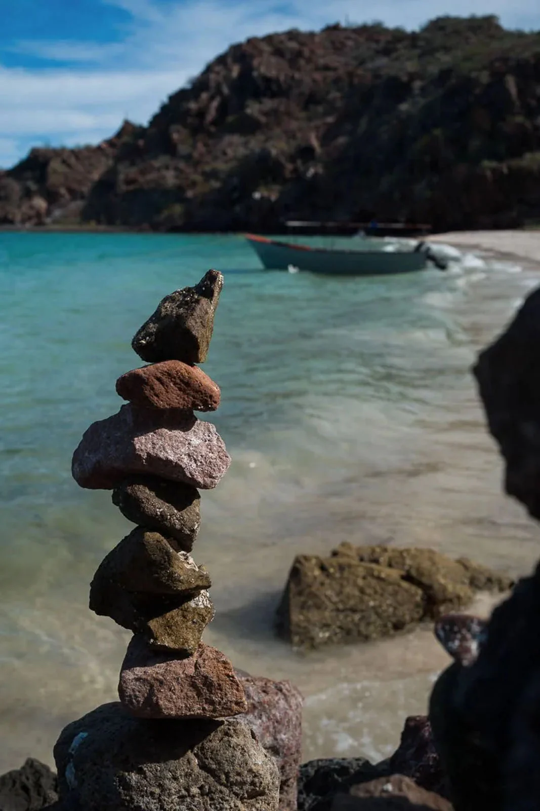 WorthWait: Fotoğrafları Görmek İçin 24 Saat Bekleten Kamera Uygulaması 20 A stack of balanced rocks stands in the foreground on a beach, with a small boat floating in the turquoise water behind it.